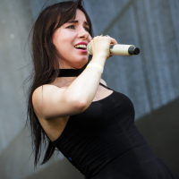 At the 2014 Piens Fest, a woman with long dark hair is passionately singing into a microphone on an outdoor stage. She is wearing a black sleeveless top and a choker necklace. Her right arm is slightly bent, and she appears to be deeply engaged in her performance. The background includes stage equipment.