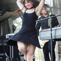 At Piens Fest 2014, a woman with long brown hair, wearing a black sleeveless dress, performs energetically on stage. She holds a microphone and has one arm lifted above her head. A man in a dark shirt plays a keyboard in the background, with stage equipment visible behind them.