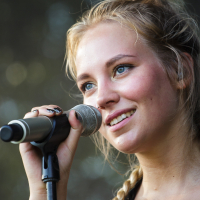 A young woman with braided blonde hair smiles as she holds a microphone near her face. She has blue eyes and wears a ring on her right hand. The background is blurred, suggesting an outdoor setting at the 2014 Piens Fest, where she appears to be enjoying the moment, possibly performing or speaking.