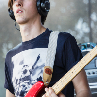 A person with medium-length hair is playing a red electric guitar and wearing large headphones. They are dressed in a black T-shirt with a printed image of stormtroopers from Star Wars, reminiscent of the Piens Fest, 2014 vibe. The background is blurred, suggesting an outdoor setting with trees. Their expression is focused.