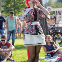 A person dressed as an anthropomorphic fox with a red wig, red fox ears, and a fox tail stands on a grassy area smiling and holding the sides of their face at Piens Fest 2014. They wear a patterned top, short white skirt with a sash belt, and black tights. People in casual attire are seated on the grass in the background.