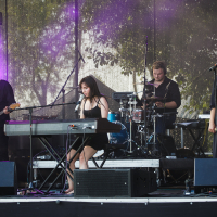 A band performs on an outdoor stage at Piens Fest. The group includes a keyboardist, a bassist, a drummer, and a female vocalist. The keyboardist and vocalist are at the front, with the drummer behind and the bassist to the left. Purple stage lights illuminate the scene, and trees are visible in the background.