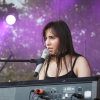 A woman with long dark hair, wearing a sleeveless black top, is singing into a white microphone while playing a keyboard on an outdoor stage at the 2014 Piens Fest. The background features purple lighting and blurred foliage. She appears focused and immersed in her performance.