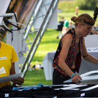 At Piens Fest 2014, two women at an outdoor event work at separate tables with various materials. The woman on the left, wearing a yellow shirt and name tag, appears to be organizing or inspecting items. The woman on the right, in a colorful vest and headband with a feather, arranges materials on her table.