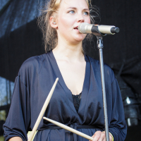 A woman with blonde hair tied back is singing into a microphone on a stand. At Piens Fest 2014, she is holding two drumsticks and wearing a dark, loose-fitting outfit with a tied waist. The background is a black stage setup. The image captures her in a performance setting.