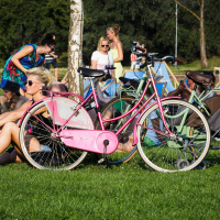 People are relaxing on a sunny day in a park. A woman is sitting on the grass near two bicycles, one pink and one green. In the background, more people are sitting and chatting, some on lounge chairs. Trees and greenery are visible, creating a serene atmosphere reminiscent of Piens Fest 2014.