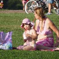 A woman and a child sit on a picnic blanket in a grassy area during Piens Fest 2014. Both are wearing pink swimwear and sun hats, drinking from straws. A transparent purple bag with items inside sits near them. Several people and bicycles are present in the background, enhancing the bright and sunny scene.