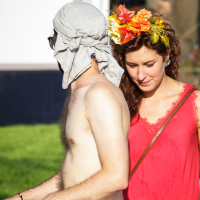 A shirtless person with a headscarf and sunglasses stands next to a woman dressed in a bright red dress and adorned with a vibrant flower crown. It's Piens Fest 2014, set outdoors on a sunny day, with grassy areas and trees in the background. The woman's gaze is downward, focused on something out of frame.