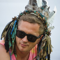 A man with short blonde hair wears black sunglasses and a headdress adorned with various colored feathers and cloth strips. He is dressed in a tie-dye tank top featuring shades of pink, yellow, and blue. The bright, blurred outdoor setting evokes memories of Piens Fest 2014.