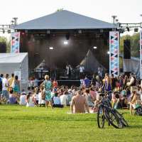 People gather at Piens Fest 2014, an outdoor music festival with a stage in the background. Some are sitting on the grass, while others stand. Bicycles are parked in the foreground. Stacks of speakers and colorful banners hang beside the stage, which is covered by a tent. Trees are visible in the distance.