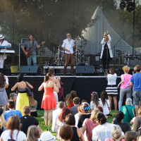 At Piens Fest 2014, a band performs live on an outdoor stage, featuring six musicians playing various instruments. An audience of diverse people stand and sit on the grass, watching enthusiastically. The stage is set against trees and is equipped with sound and lighting equipment, enhancing the lively atmosphere.