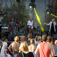At Piens Fest 2014, a band performs on an outdoor stage with a drum set, keyboard, electric guitar, and bassist. Two vocalists sing at center stage, one dressed in a long jacket. Audience members, many in summer attire, watch and dance as sunlight filters through the trees, illuminating the scene.