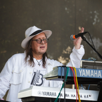 A person wearing a white outfit and a white fedora hat plays a Yamaha keyboard on stage at Piens Fest 2014. They have long hair and wear glasses, appearing focused on the music. A microphone is positioned in front of them, and colorful cables connect to the keyboard. Tree branches are visible in the background.