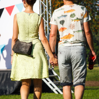 A couple is walking hand in hand in an outdoor setting during Piens Fest 2014. The woman is wearing a light green dress, sunglasses, and a black crossbody bag. The man is dressed in a white t-shirt with a colorful print and gray shorts, holding a red bottle. They are seen from behind on a sunny day.