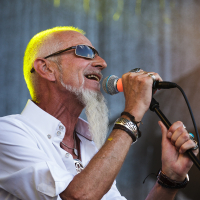An older man with a white beard, sunglasses, and a short-sleeved white shirt sings passionately into a microphone at the 2014 Piens Fest. He wears various bracelets, rings, and a necklace. The background, slightly out of focus, includes a mesh-like backdrop and yellow stage lighting.