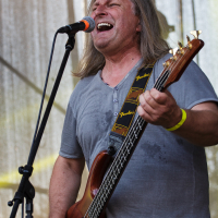 A man with long gray hair passionately sings into a microphone while playing a brown electric bass guitar on stage at Piens Fest 2014. He is wearing a gray short-sleeve shirt and a yellow wristband, with a "Fender" brand strap visible. The background features a mesh screen with stage equipment partially visible.