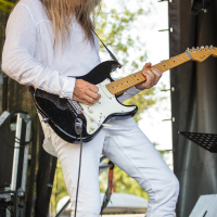 A person with long hair, wearing an all-white outfit, is playing an electric guitar outdoors. The individual is strumming a black and white guitar with detailed focus. The background features stage equipment and a black backdrop. Trees and an audience can be seen in the distance at Piens Fest 2014.