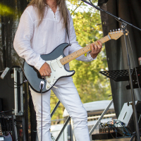At Piens Fest 2014, a man with long hair and glasses passionately plays an electric guitar and sings into a microphone on stage. He is dressed in white from head to toe, wearing a long-sleeve shirt, pants, and sneakers. Behind him, stage equipment and trees are visible.