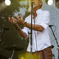 An elderly man with a white beard and sunglasses passionately sings into a microphone on the Piens Fest stage. He wears a white button-up shirt with rolled sleeves and brown pants, accessorized with rings, bracelets, and a necklace. Bright stage lights illuminate him while drum sets are visible in the background.