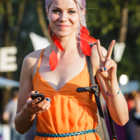 A woman with purple hair, wearing an orange dress, holds a phone and makes a peace sign with her right hand. She is accessorized with red feather earrings and a blue belt. The background is outdoors at the lively 2014 Piens Fest, filled with blurred greenery and people enjoying the festivities.