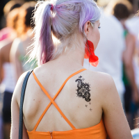 A woman with light purple hair pulled into a ponytail stands in a crowd at Piens Fest 2014. She is wearing a bright orange dress with thin straps crossing over her back. On her left shoulder blade is a large, intricate black henna tattoo. Her left earring is large and red. She has a black strap over her shoulder.