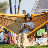 A boy wearing a gray T-shirt, beige pants, and a bucket hat with stars sits in a yellow hammock while looking at his phone. He's outdoors at Piens Fest 2014. The background shows people sitting on the grass—some relaxing and some talking—with a tree and tents visible behind them.