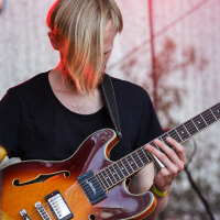 A person with blond hair, dressed in a black shirt, is playing an amber-colored electric guitar on stage at Piens Fest 2014. Focused on the instrument, the guitar strap sits over their shoulder. The background is a textured, light-colored curtain under red stage lighting.