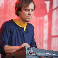 A person with medium-length brown hair, wearing a blue shirt with yellow detail at the collar, intensely plays a keyboard adorned with "Rogue" and "Moog" labels. The red and blurred background, coupled with an indistinct shadowy figure, suggests this could be a live performance at Piens Fest 2014.
