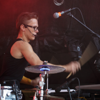 A musician with short hair and glasses is energetically playing the drums on stage at Piens Fest 2014. They are wearing a sleeveless black shirt. The scene is under red lighting with a microphone positioned near their mouth, suggesting they might be singing. Various drum equipment and wires are visible around them.