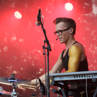 A drummer wearing a black tank top and glasses performs passionately on stage under red lighting at the 2014 Piens Fest. With two cymbals and an electronic keyboard setup, he plays with intensity, a microphone positioned in front of him. The blurred background keeps the focus on the musician.