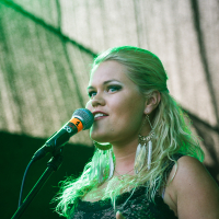A woman with long blonde hair, partially tied back, stands on stage at the 2014 Fest, holding a microphone. She is wearing a black lace top and large feather earrings. The background is dark with green lighting. She appears to be singing or speaking.