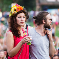 A woman wearing a floral headpiece and a pink dress holds a clear cup while standing beside a man with a beard, sunglasses, and a gray T-shirt. The man is looking to his left with a cigarette in his mouth. The background shows people sitting and standing on the grassy area of Piens Fest 2014, indicating an outdoor event.