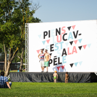 Two people stand on an outdoor stage beside a large sign with colorful, geometric text at Piens Fest 2014. They pose for a photo on a sunny day in the park. Several people sit or lie on the grass, enjoying the open, grassy field and warm weather. Trees and playground equipment are visible in the background.