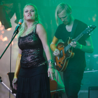 A blonde woman in a black lace top and skirt sings passionately into a microphone on stage at Piens Fest 2014. Beside her, a man with long blonde hair plays an electric guitar. The stage background is green, with blurred lights hinting at an outdoor night setting. Musicians and equipment are in the background.