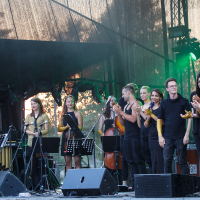 At Piens Fest 2014, a group of musicians and singers stand on an outdoor stage, dressed in black attire, holding various instruments, including violins and a cello. They are smiling and clapping, illuminated by stage lights, with a backdrop of trees and a dark canopy overhead. Microphones and speakers are visible.