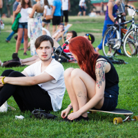 Two individuals sit on grass at an outdoor gathering during Piens Fest 2014. The person on the left wears a white t-shirt and black pants, while the person on the right has red hair, a black tank top, shorts, and a visible tattoo. A skateboard lies beside them. In the background, multiple people interact and bicycles are visible.