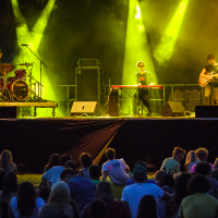 A live band performs on stage with dramatic yellow lighting at Piens Fest 2014. The drummer is on the left, a keyboard player in the middle, and a guitarist on the right. An audience sits on the grass, watching intently. Smoke effects and amplifiers are visible in the background.