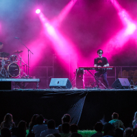 A live music performance on an outdoor stage with three musicians at Piens Fest 2014. From left to right, a drummer, a keyboardist, and a guitarist play under vibrant pink and purple stage lights, producing a misty, atmospheric effect. An audience watches attentively from the grass in front of the stage.