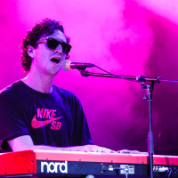 A person with curly hair, wearing sunglasses and a black Nike SB shirt, sings into a microphone while playing a red and white Nord keyboard on stage at Piens Fest 2014. The background is lit with purple and pink lights, creating a vibrant atmosphere.