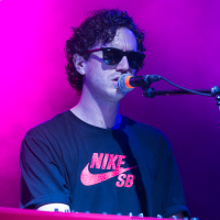 A person with curly hair and sunglasses is playing a red Nord keyboard at Piens Fest 2014. They are wearing a black Nike SB t-shirt and singing into a microphone attached to a stand. The background is illuminated with purple and pink stage lights, creating a vibrant atmosphere.