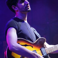 A musician with short dark hair and facial hair is playing an electric guitar on stage at Piens Fest 2014. He is wearing a black t-shirt and is illuminated by purple stage lighting, with a pensive expression. The guitar has a sunburst finish. The background is dark and out of focus.