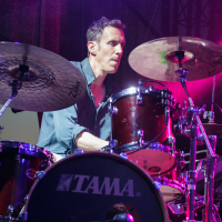A drummer is intensely playing a TAMA drum set on stage at Piens Fest 2014. The stage is illuminated with colorful lighting, primarily purple, creating a dynamic atmosphere. He is surrounded by cymbals and drum pieces, wearing a long-sleeved shirt with rolled-up sleeves, focused on his performance.