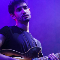 At Piens Fest 2014, a man wearing a black T-shirt plays an electric guitar on stage. He has short curly hair and a beard. The background is illuminated with purple lighting, creating a vibrant atmosphere. The man looks slightly upwards, engrossed in the music he is playing.