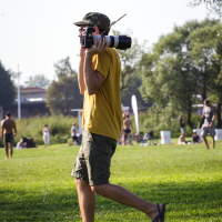 A person dressed in a yellow t-shirt, camouflage hat, shorts, and flip-flops is walking on a grassy field at Piens Fest 2014. They are carrying a camera with a long telephoto lens on their shoulder. The background shows other people relaxing and enjoying a sunny day in the park with trees and blue sky.