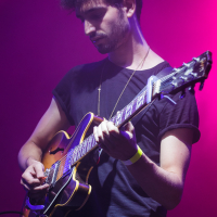 At Piens Fest 2014, a guitarist with dark, curly hair and a trimmed beard stands on stage, playing an electric guitar. He wears a black t-shirt and a yellow wristband on his right arm. The background is illuminated with purple and pink stage lights, creating a vibrant atmosphere.
