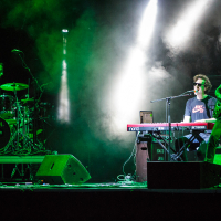 At Piens Fest 2014, a three-member band performs on stage. The drummer is playing a drum set on the left, the keyboardist is in the center singing into a microphone, and the guitarist stands on the right. Green and white stage lights and fog create a dramatic atmosphere. The drum kit has "TAMA" written on it.