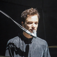 A person with short, dark hair, wearing a dark shirt, is standing behind a microphone and a drum set. They are looking focused, likely performing on stage at Piens Fest 2014, with dramatic lighting casting shadows on their face.