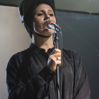 A person wearing a dark, hooded outfit sings into a microphone with a red pop filter. They have short hair and are standing in front of a dimly lit, blurred background. The expression on their face suggests they are deeply engaged in the performance, evoking memories of Piens Fest 2014.