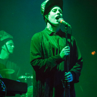 A singer in a dark costume and headpiece performs onstage under green lighting at Piens Fest 2014, gripping a microphone with both hands. A drummer in similar attire is visible in the background, playing on a drum set. The scene conveys a dark, moody atmosphere, enhanced by the stage lighting.