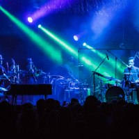 At Piens Fest 2014, a live music performance unfolded on a dimly lit stage. A pianist and drummer took the foreground while musicians playing brass instruments stood behind them, surrounded by sound equipment. Vibrant blue and green lights illuminated the stage as the captivated crowd watched.